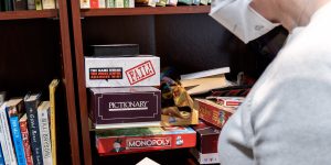 A person with short hair and an N95 mask standing in front of some bookshelves labeled "Recovery Books" reading a book
