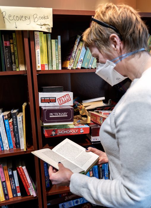 A person with short hair and an N95 mask standing in front of some bookshelves labeled "Recovery Books" reading a book