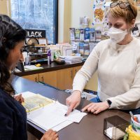Person checking in at the front desk of a recovery support organization listens while a staff person points at a ledger and explains the process