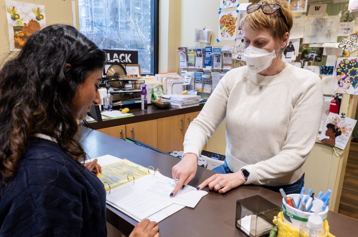 Person checking in at the front desk of a recovery support organization listens while a staff person points at a ledger and explains the process