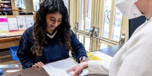 Person checking in at the front desk of a recovery support organization looks on as a staff person points at a ledger and explains the process