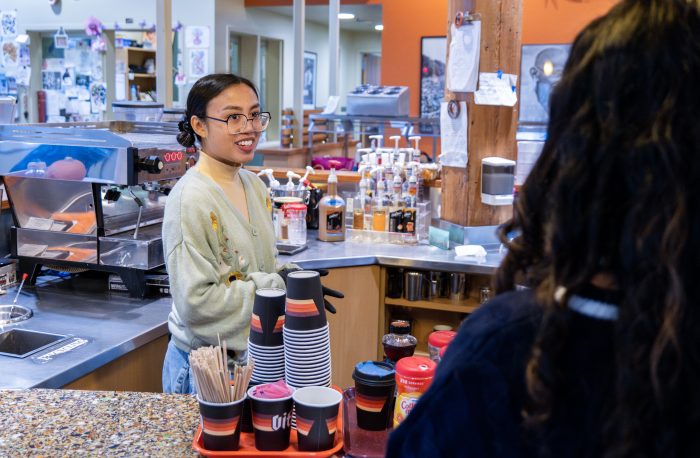 A barista with glasses and a beige sweater is smiling at someone on the other side of a cafe counter, as though taking their coffee order.
