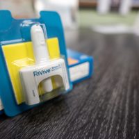 A single-use naloxone nasal spray atomizer in yellow and blue packaging sitting up against the box it came in on a wooden table.