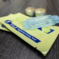 Two packages of fentanyl test strips sitting on a table with a couple of small vials of water and some small cookers/bowls in the background that can be used to combine a sample of a substance with the water for dipping the test strips in.