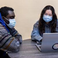 A client with a blue hoodie and a blue surgical mask sits at a table with a provider or peer support specialist, also masked. The provider/peer is showing them something on a laptop as they talk.