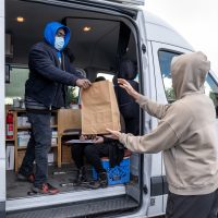 A client wearing a tan hoodie reaches out to take a brown paper bag of safer use supplies from a staff person in a syringe services program’s mobile services van. The staff person is wearing a blue surgical mask.