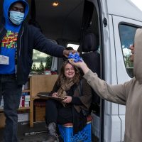 A staff person of a syringe services program’s mobile services van wearing a blue surgical mask hands some blue latex gloves from a box to a smiling client wearing a tan hoodie.